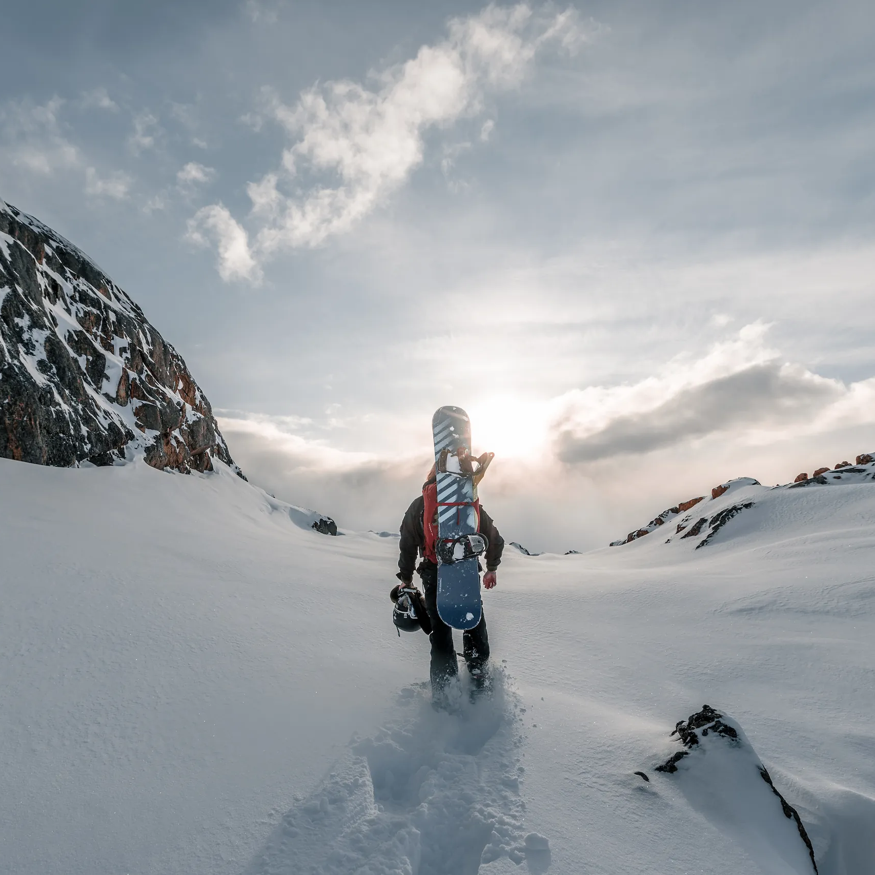 Snowboarder hoda kroz planinski sneg noseći dasku prema sunčevoj svetlosti